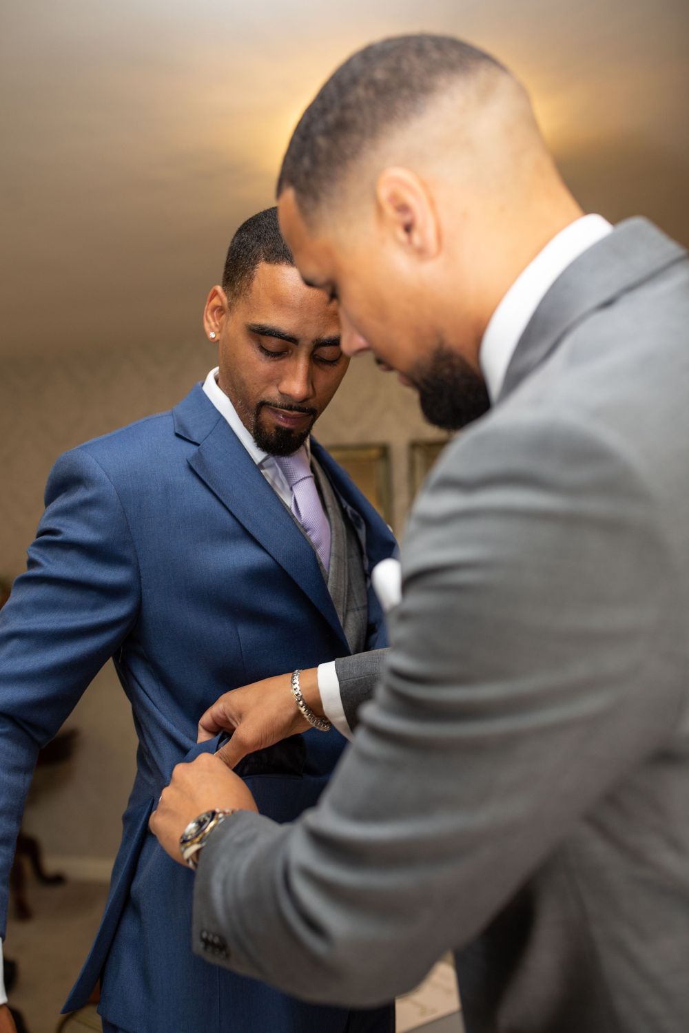 Two groomsmen help each other get dressed, one wearing a navy suit and the other in gray.
