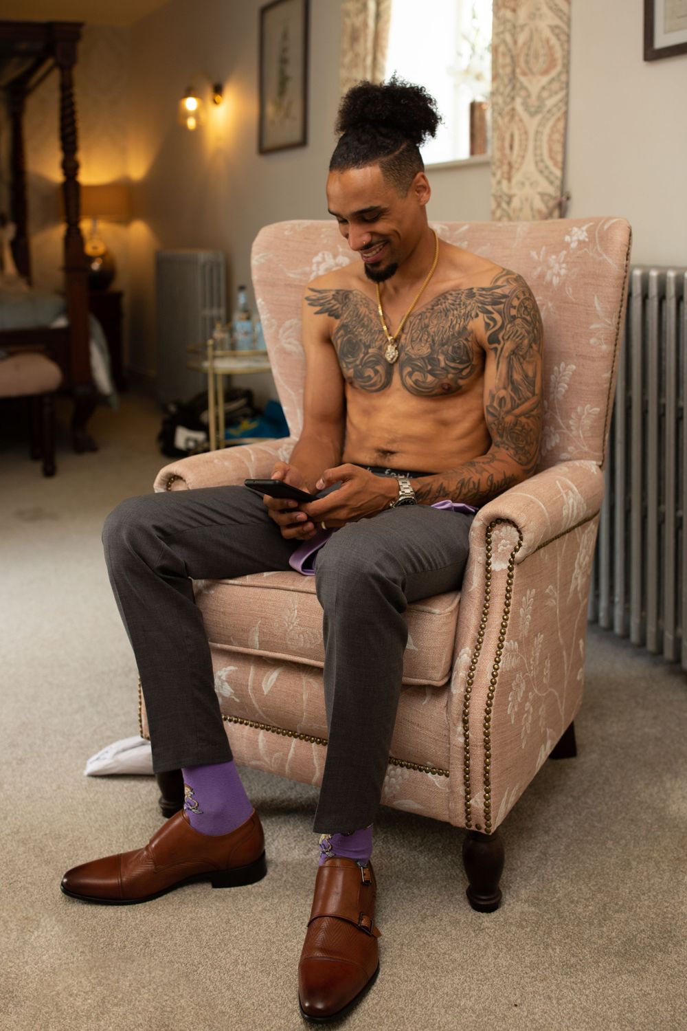 Tattooed person relaxing in vintage armchair wearing formal attire with purple socks.