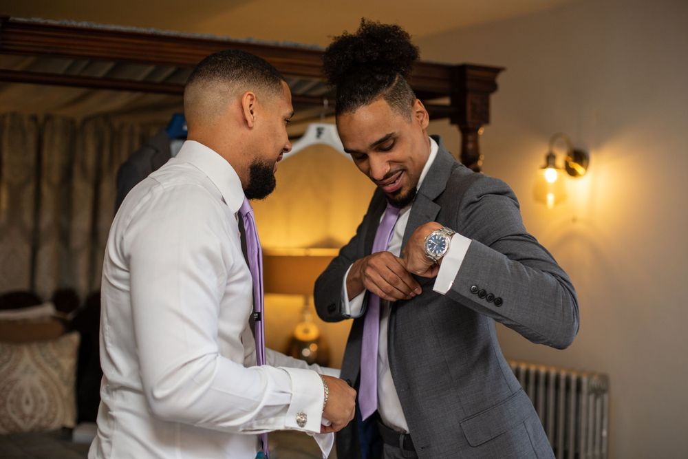 Two people in formal wear helping each other with cufflinks in dimly lit room.