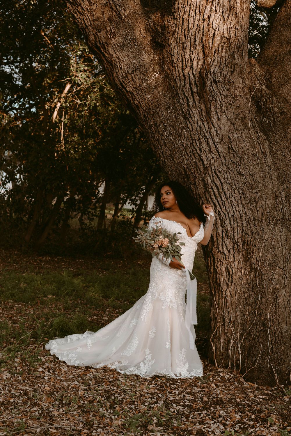 Bride in an off-shoulder lace gown holds bouquet while standing beside massive tree.