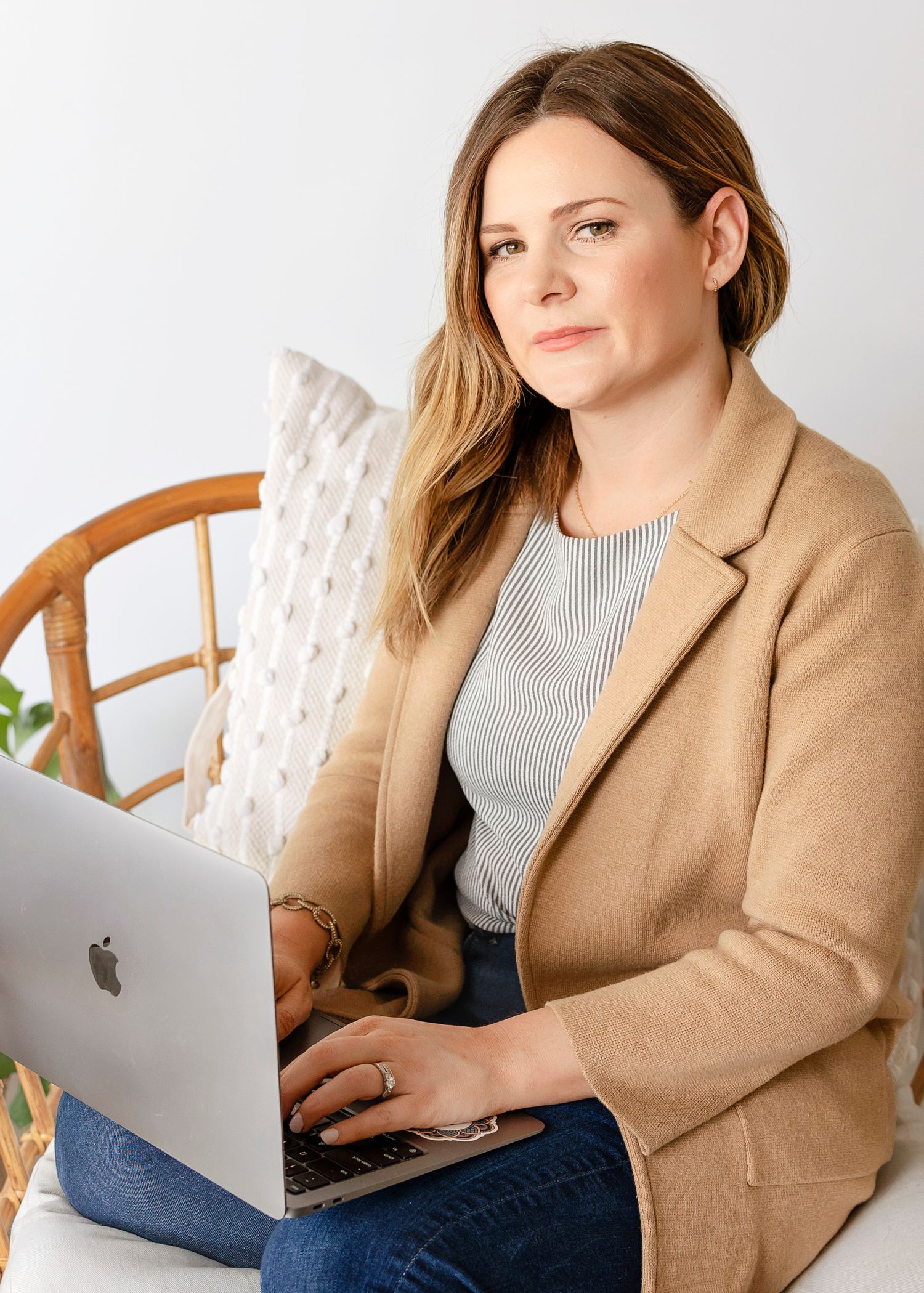 Person working on MacBook wearing tan blazer and striped top in home office setting.