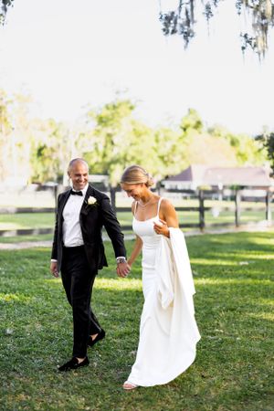 A joyful newlywed couple walks hand in hand across a sunlit lawn with Spanish moss trees overhead.