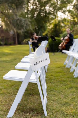 White folding chairs set up for an outdoor wedding ceremony with musicians in the background.