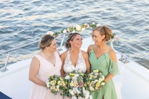 Three women in wedding attire smiling and laughing on bow of boat with floral bouquets.