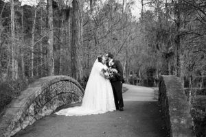 Black and white photo of newlyweds embracing on stone bridge in wooded setting.