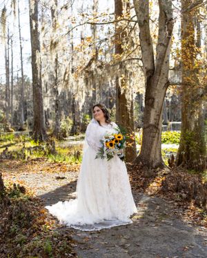 A bride in a lace gown holds a sunflower bouquet in a wooded forest setting.