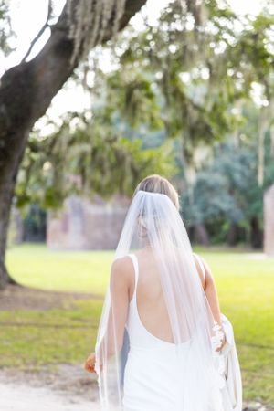 A bride in a white wedding dress with flowing veil stands under Spanish moss draped trees.
