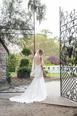 A bride in a flowing white gown stands by ornate iron gates in a garden setting.