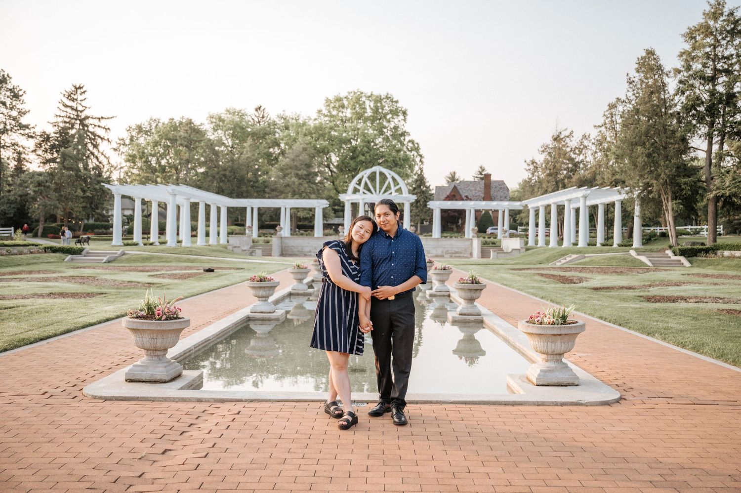 Couple poses in front of elegant white pillars and fountain in formal garden setting.