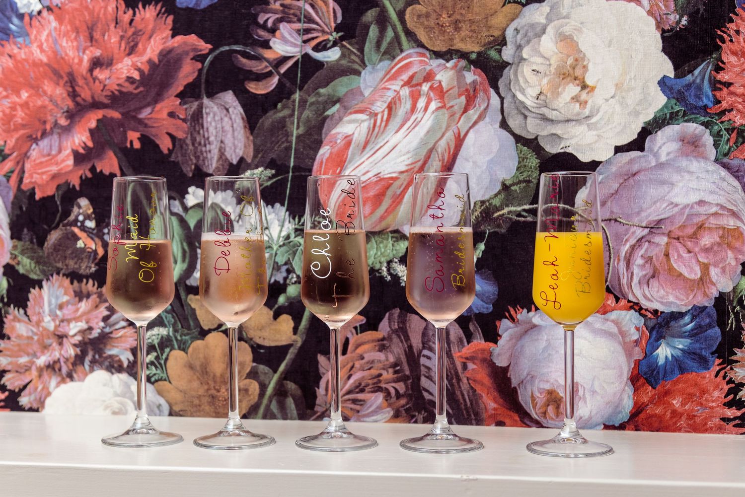 Row of champagne and mimosa glasses against a bold floral wallpaper background.