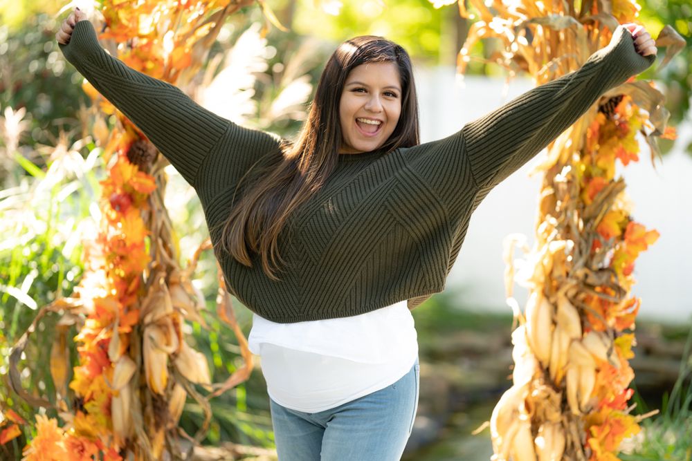 Joyful woman in dark sweater spreads arms wide while posing against autumn foliage.