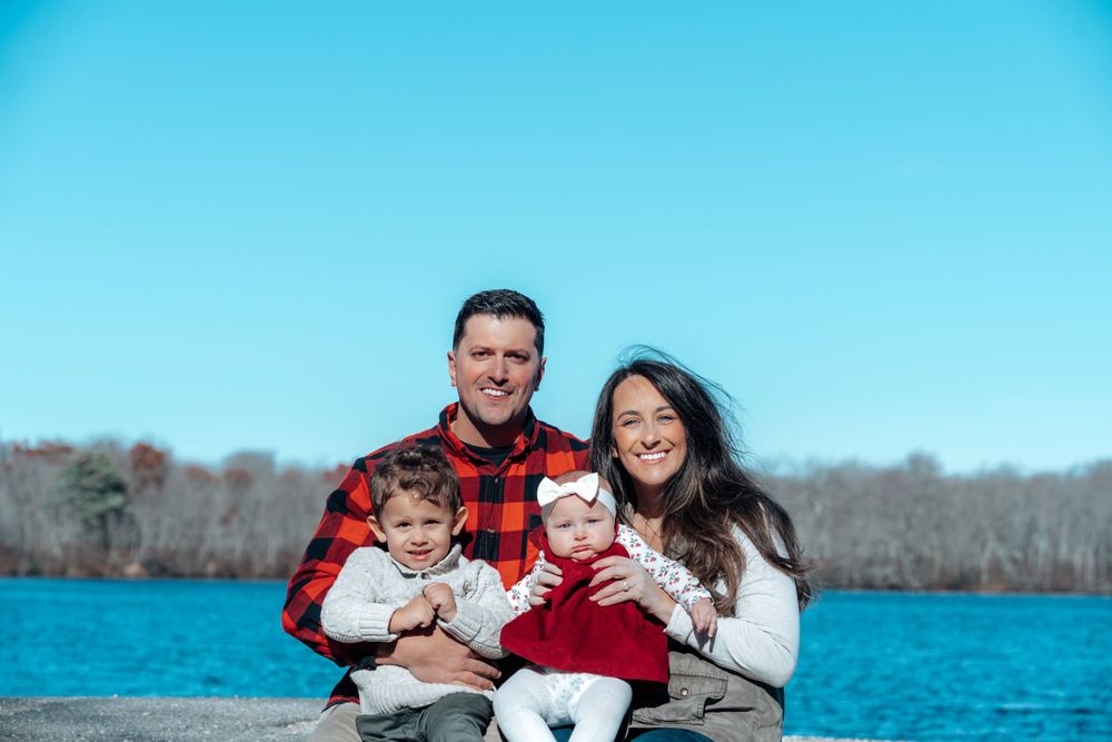 Family sits together by a bright blue lake on a winter day wearing red plaid and white sweaters.