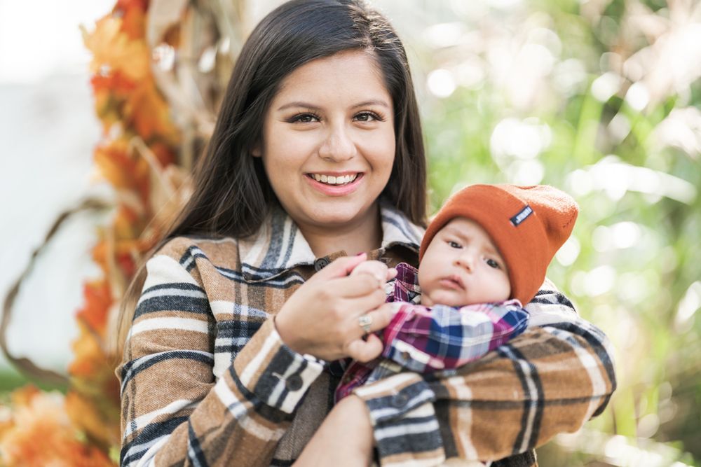Mother in plaid jacket holds smiling baby wearing orange beanie hat outdoors.