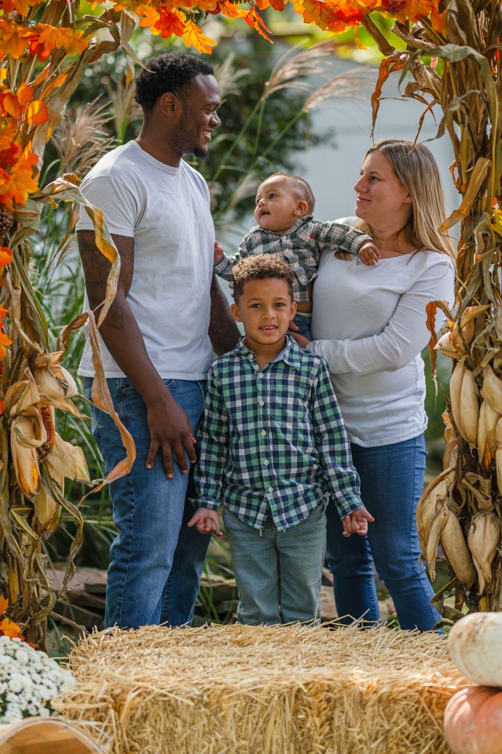 Family poses together in front of autumn corn stalks and hay bales at a fall harvest display.