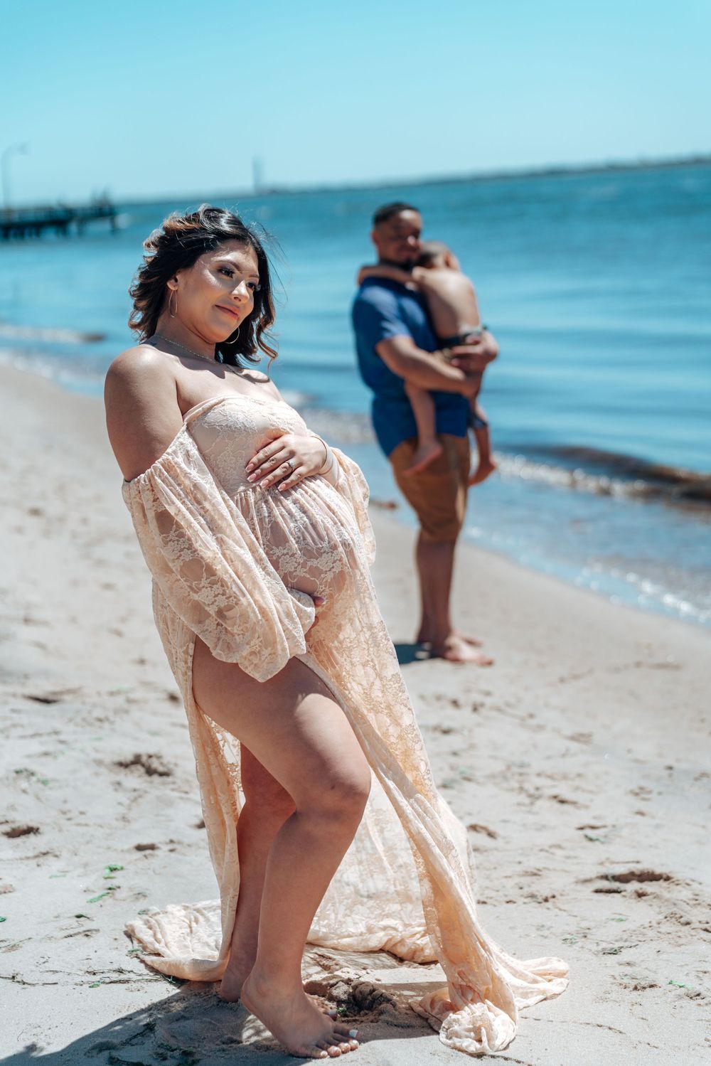 Maternity photoshoot on a beach with flowing beige dress and ocean backdrop.