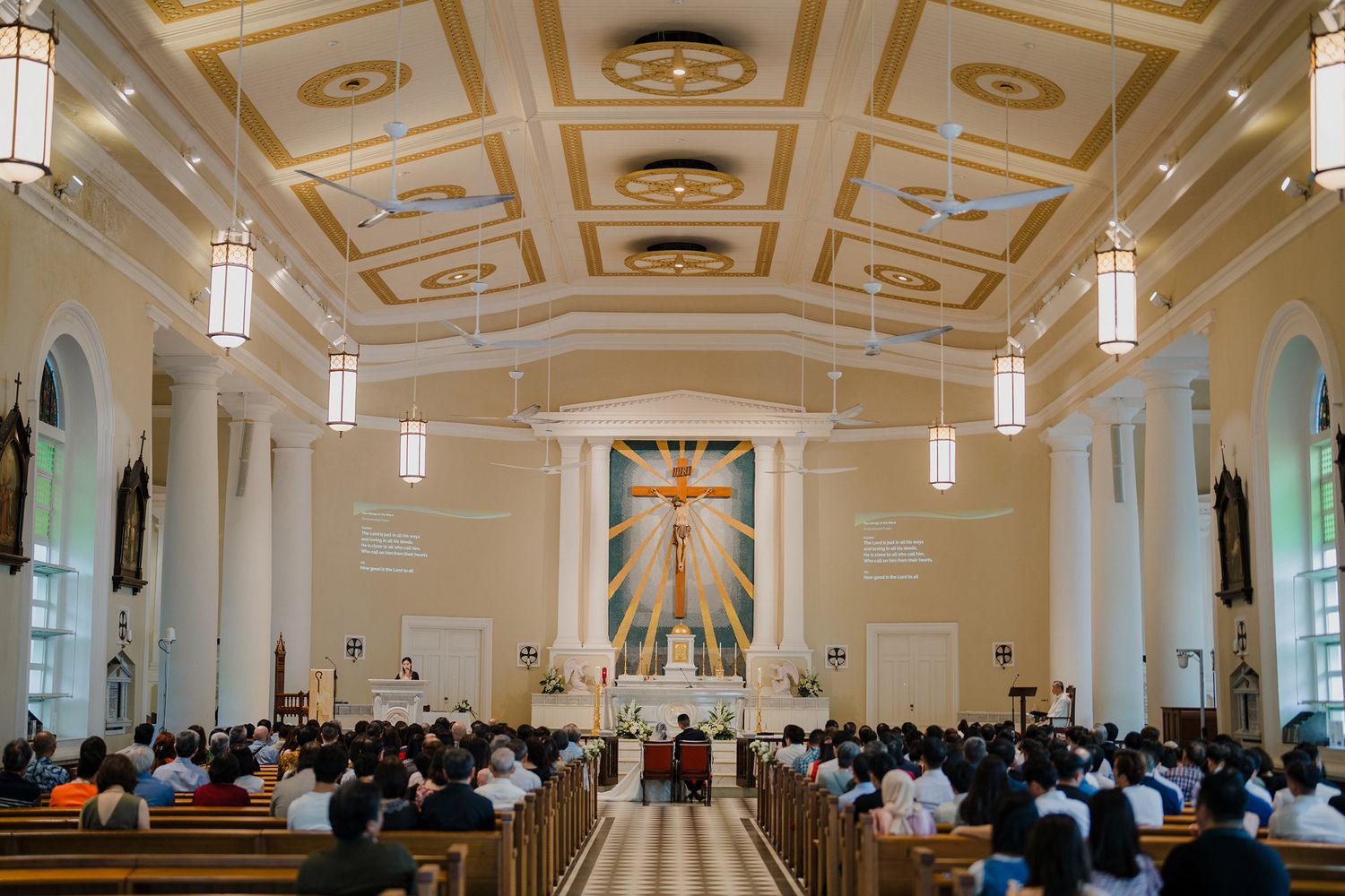 Cathedral-style church interior with ornate ceiling details and hanging pendant lights during a wedding.