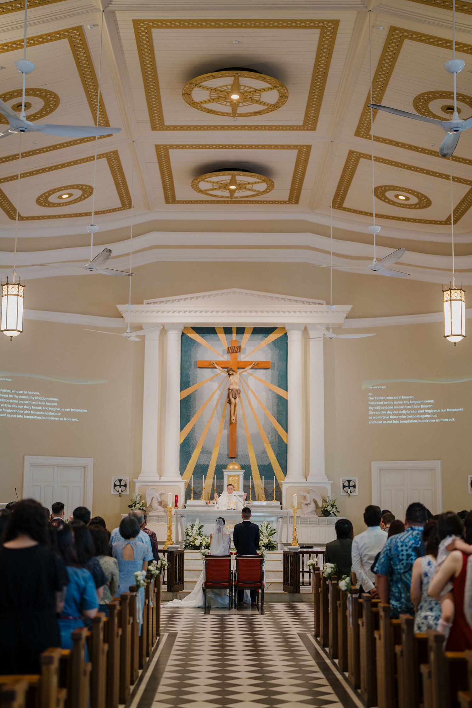 Interior of a Catholic church with decorative ceiling, pendant lights, and dramatic sunburst altar design.