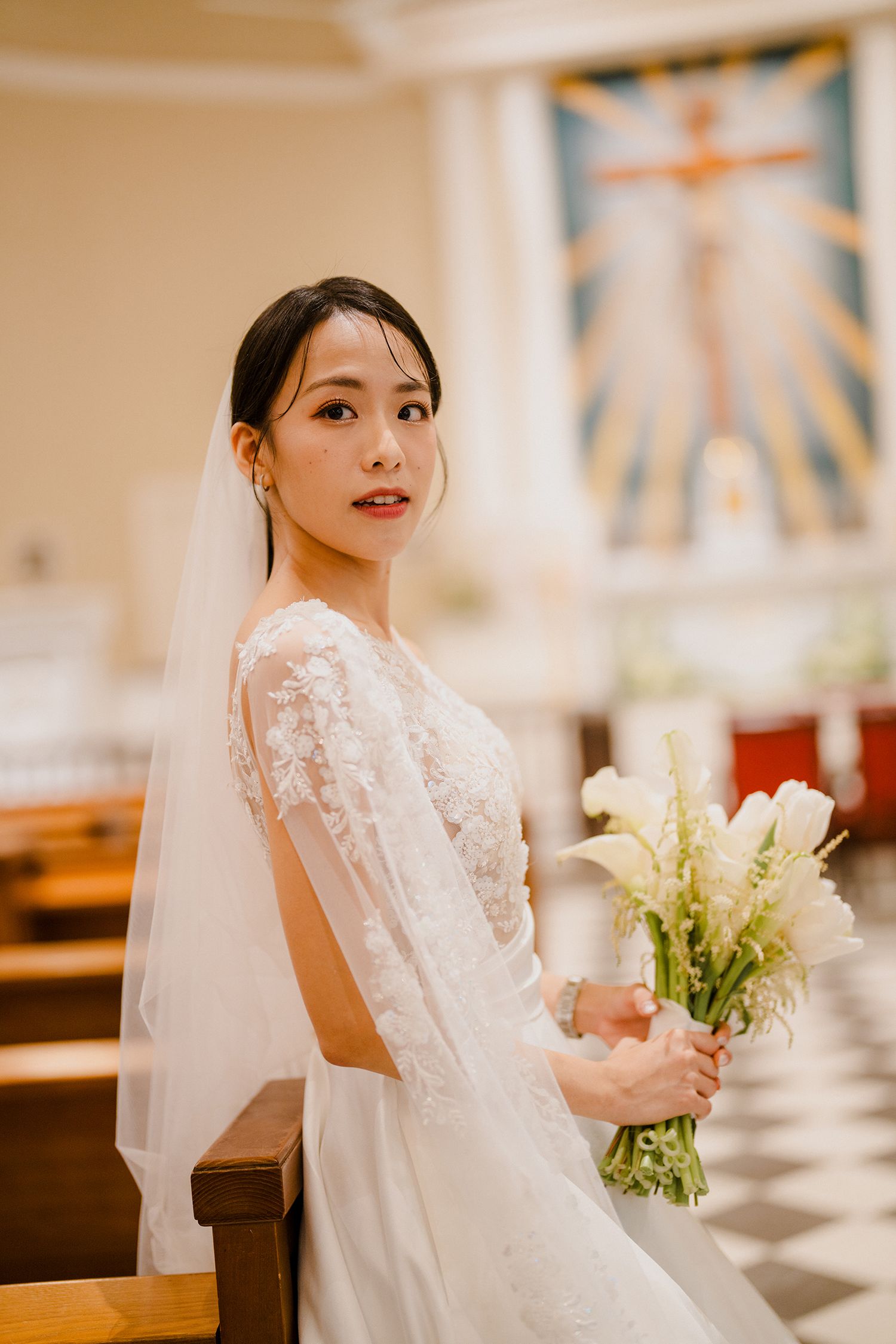 Bride in lace wedding gown holds white bouquet in church sanctuary.