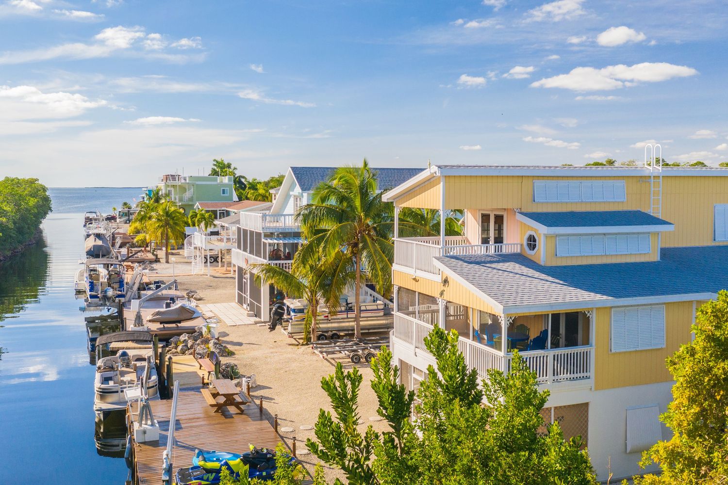 Aerial panorama of waterfront homes and palm trees along a tropical coastline with turquoise waters and boats.
