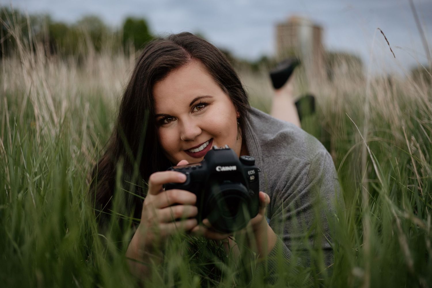 Photographer lying in tall grass field taking photos with Canon DSLR camera.