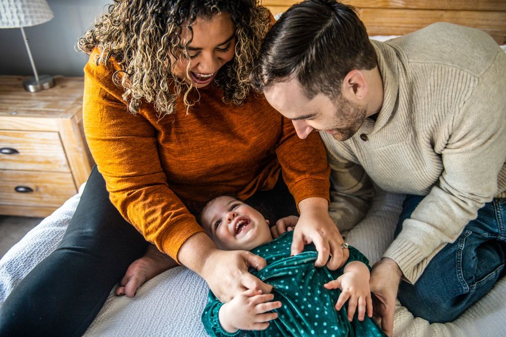 Playful family moment with parents tickling child on bed during casual weekend.