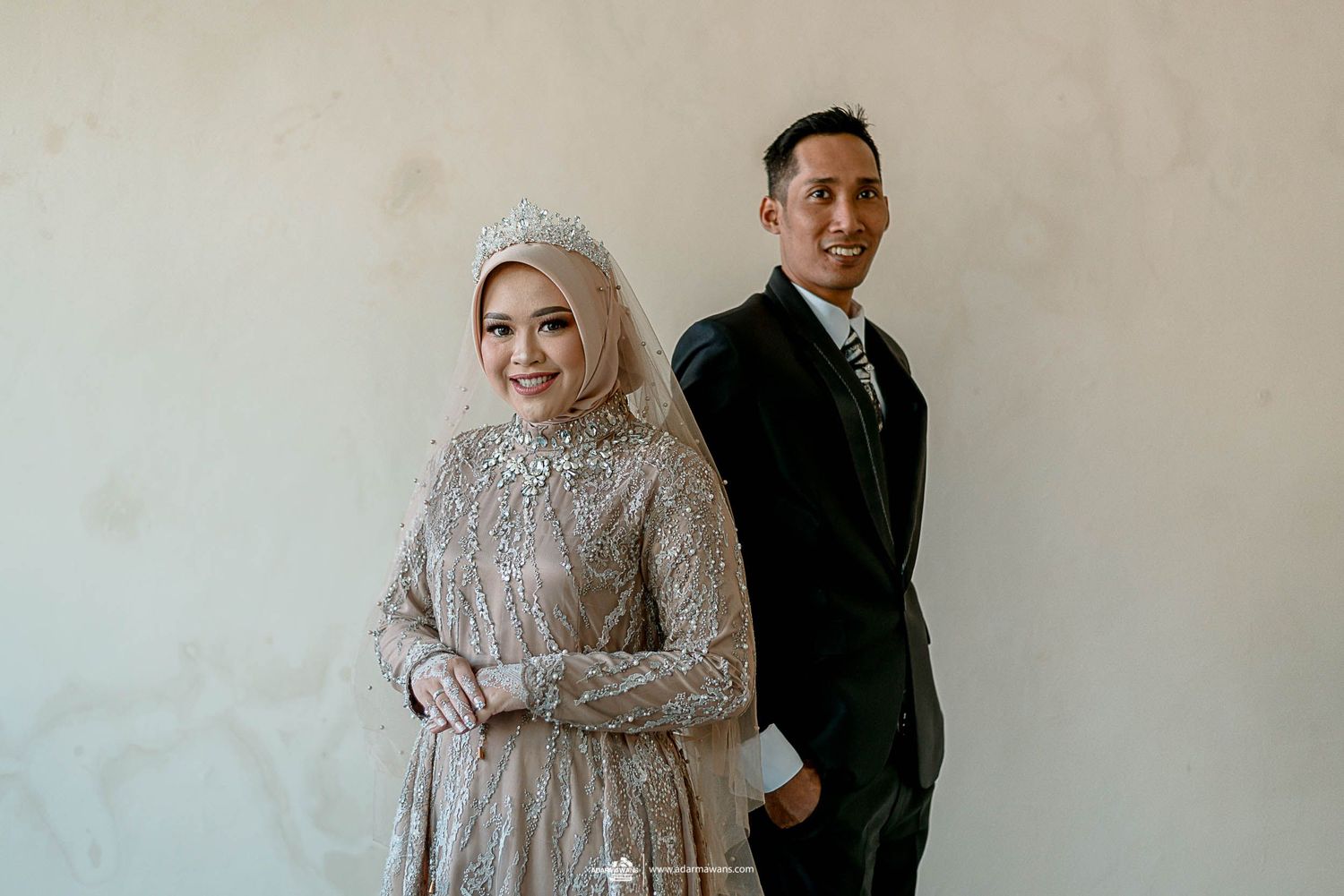 A couple poses formally against a white wall in elegant wedding attire.