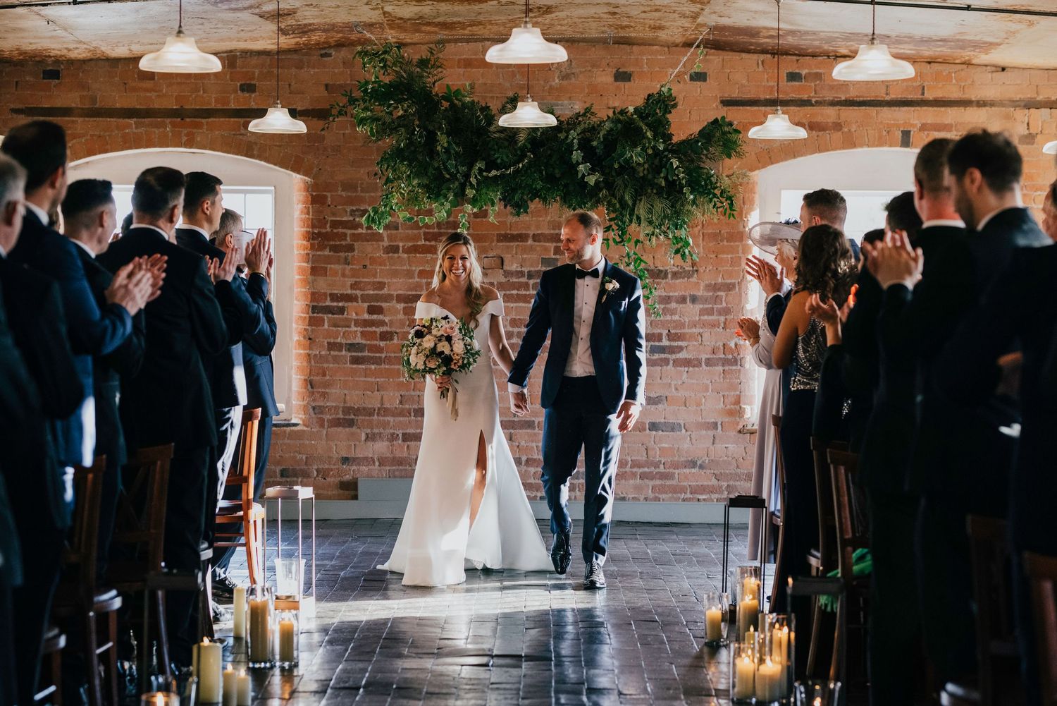 Wedding ceremony in industrial brick venue with pendant lights and aisle lined with candles.