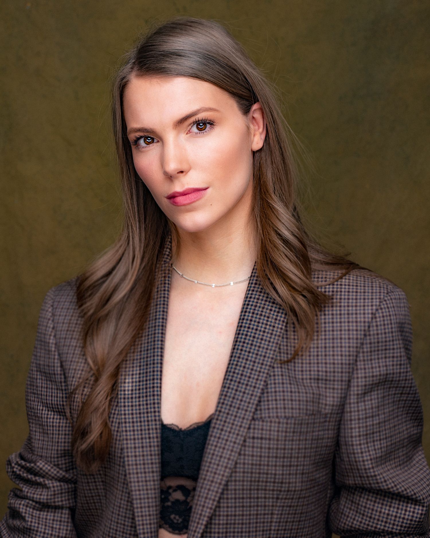 Professional headshot in plaid blazer and black top with long brown hair against olive background.