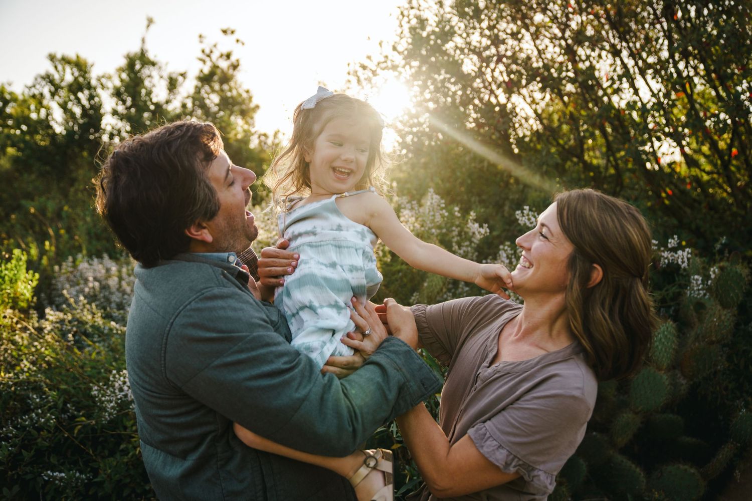 Parents playfully lifting their young child in the air during golden hour in a garden setting.