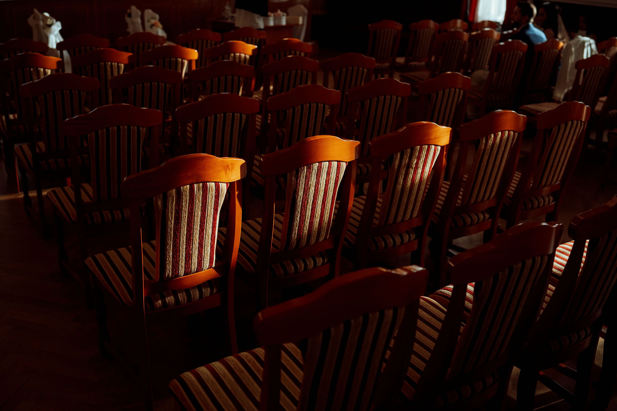 Empty wooden chairs arranged in rows within dimly lit auditorium space.