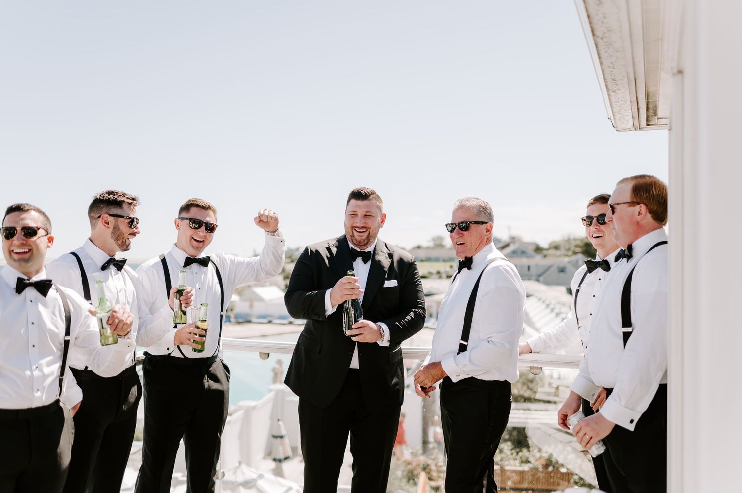 Wedding party in formal attire gathers on sunny balcony with coastal views.