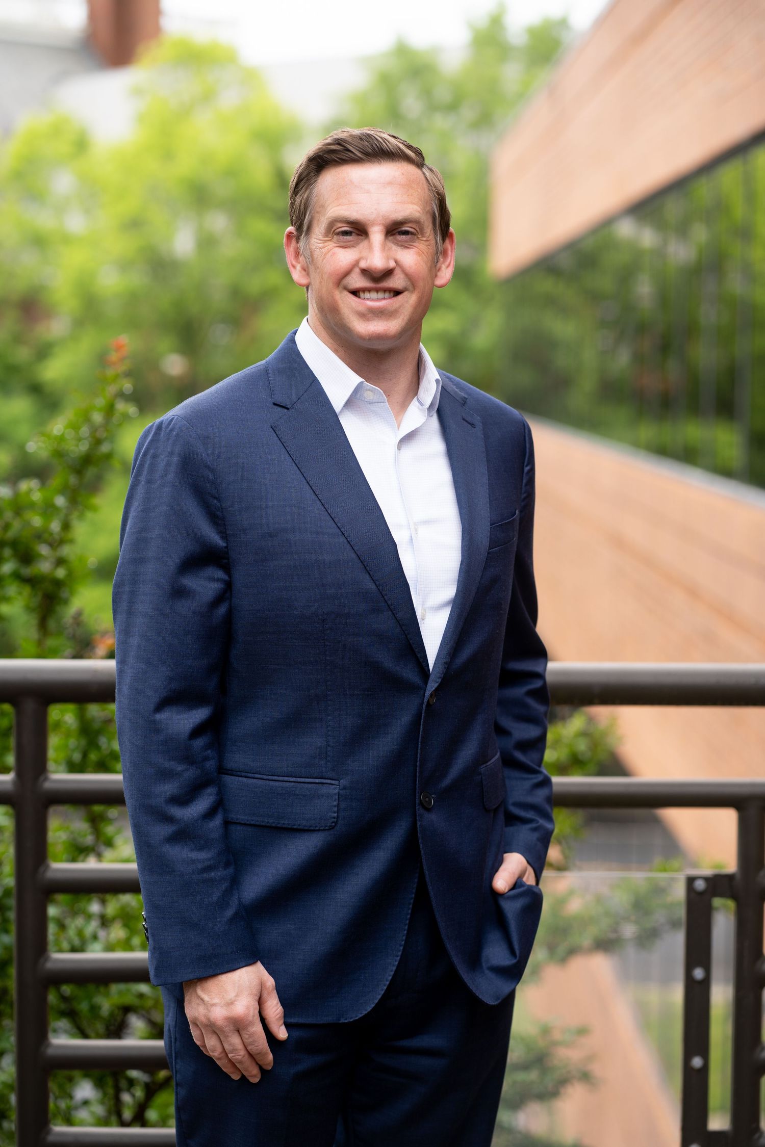 Professional in navy suit jacket standing outdoors on balcony with greenery in background.