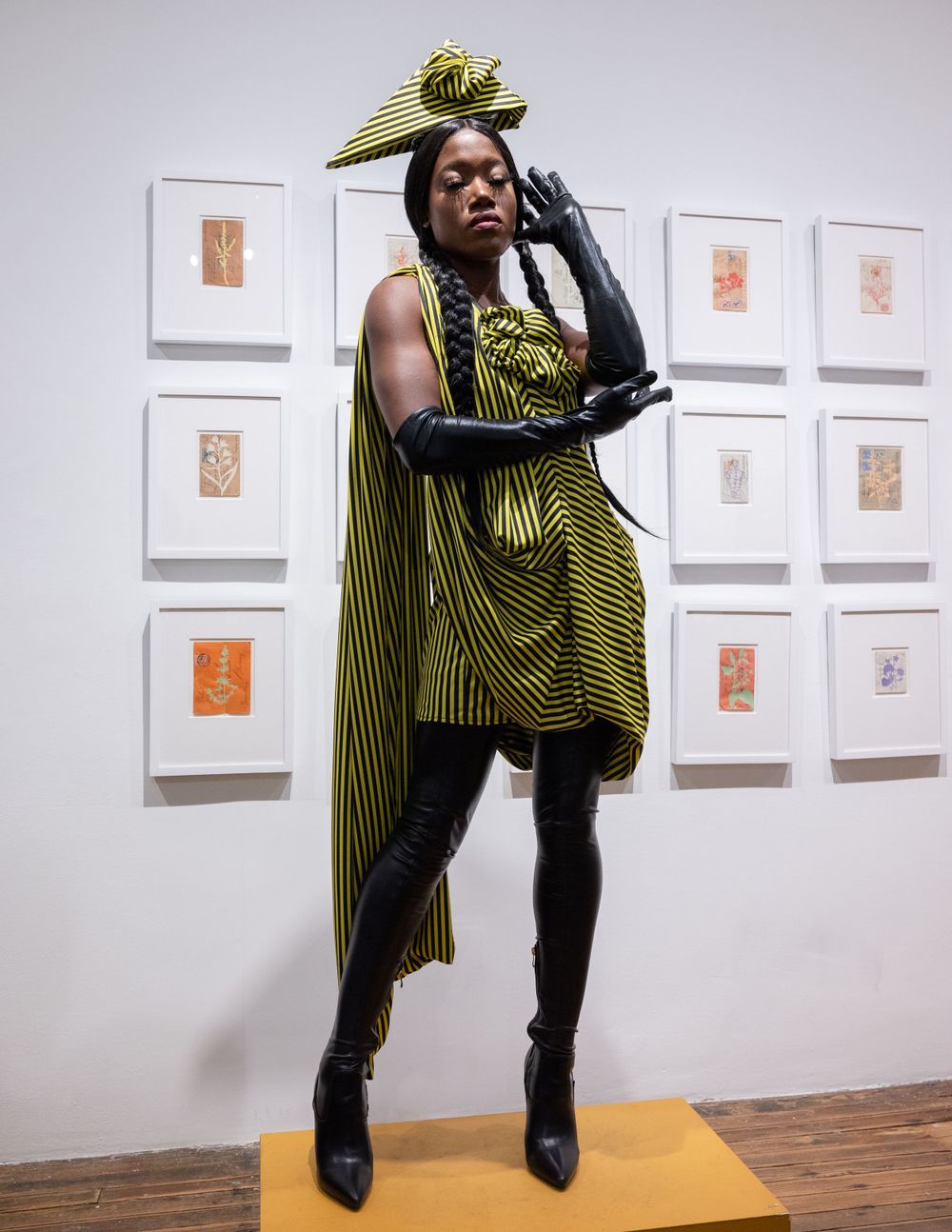Model in green striped gown with black gloves strikes dramatic pose against gallery wall with framed artwork.