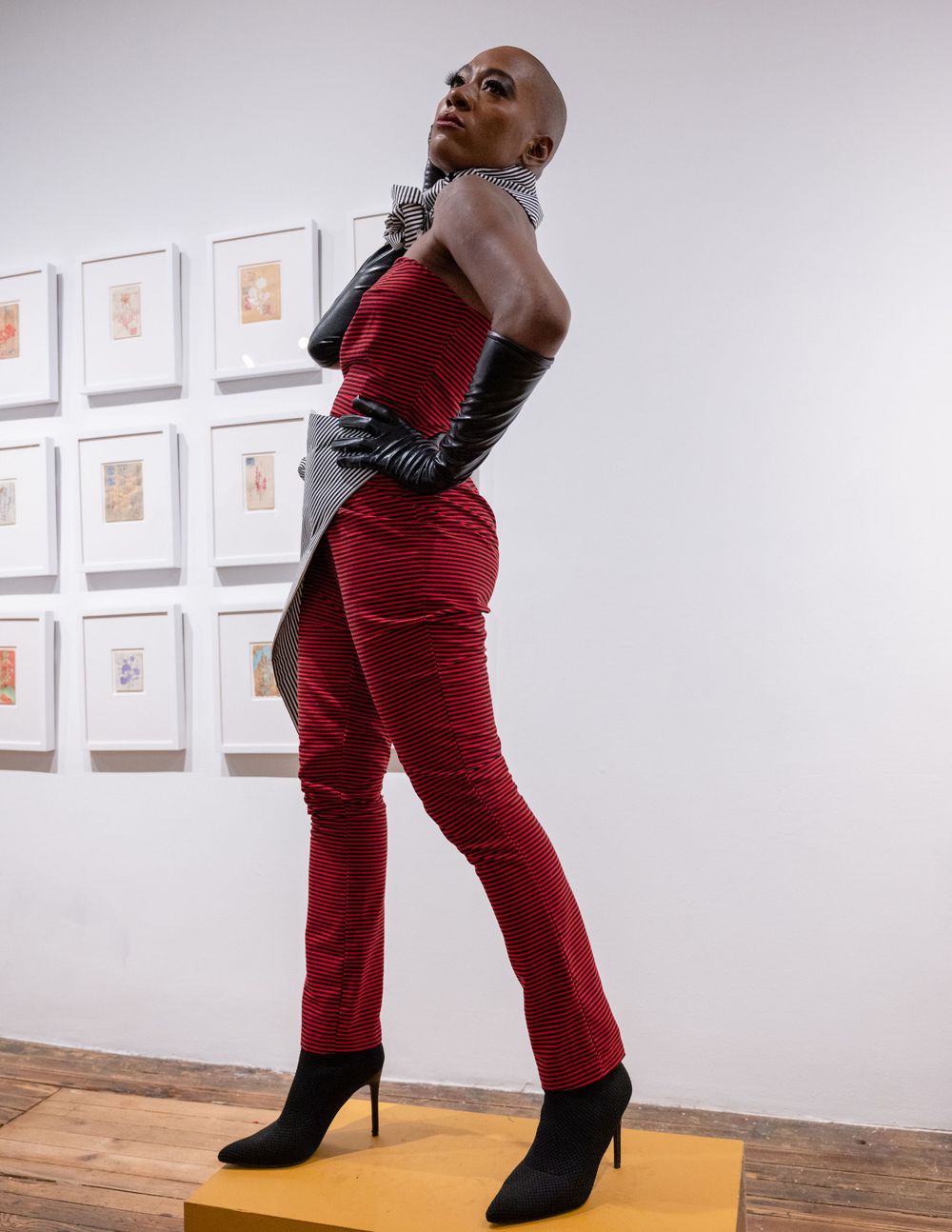 Model wearing red textured jumpsuit and black gloves poses confidently in art gallery setting.