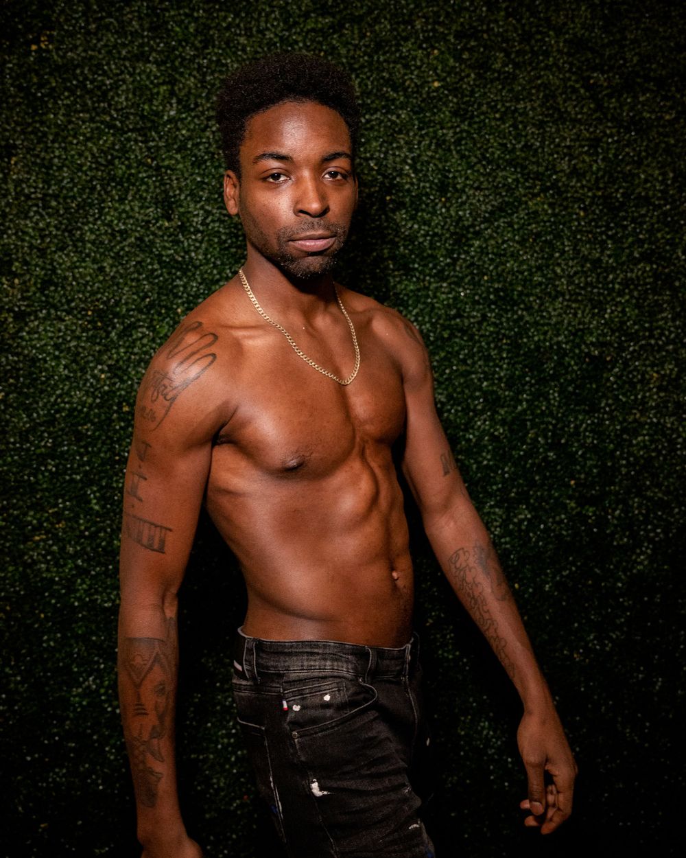 Artistic portrait against green hedge wall showing muscular physique and tattoos.