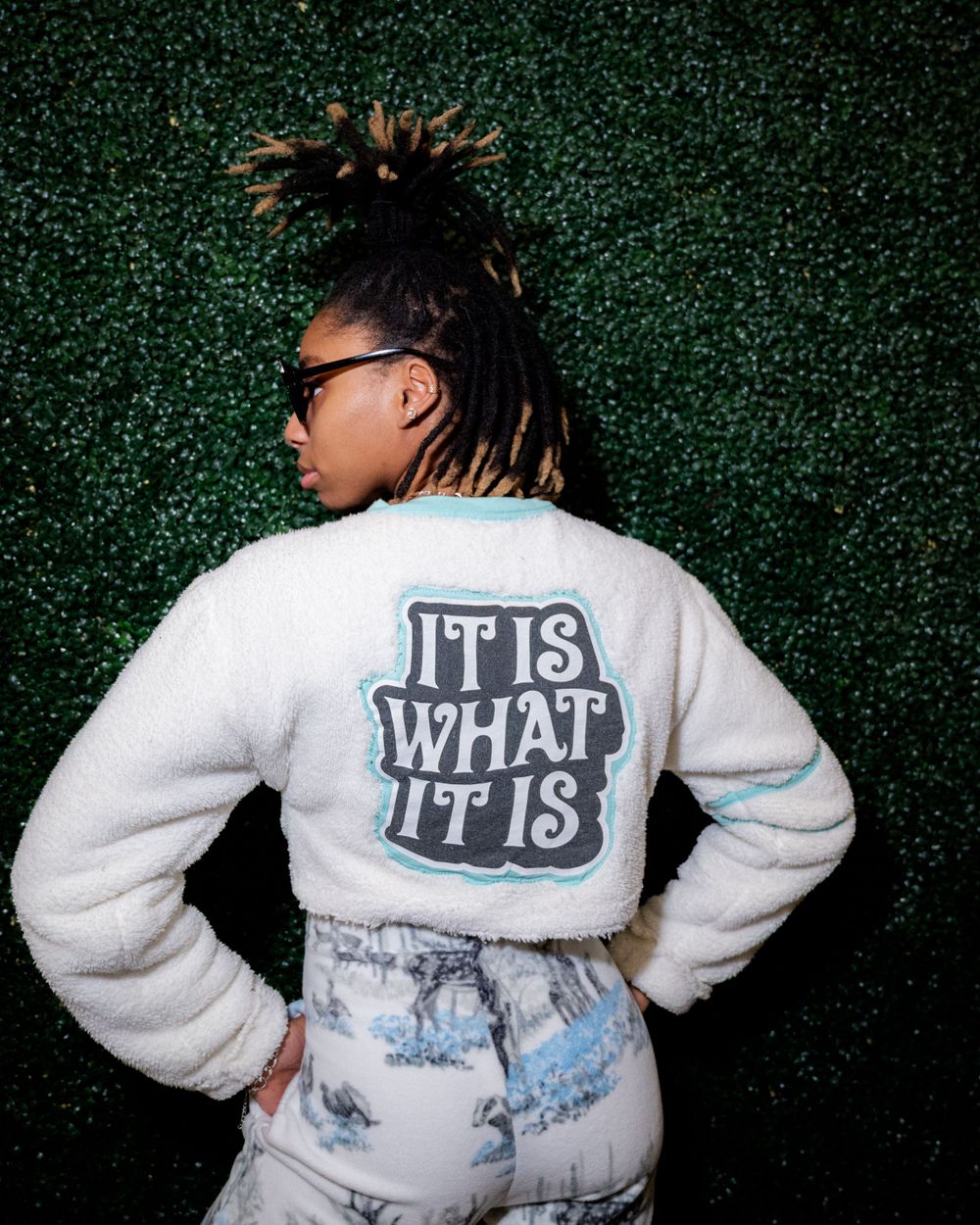 White sweater with "It Is What It Is" text shown from back against green hedge wall.