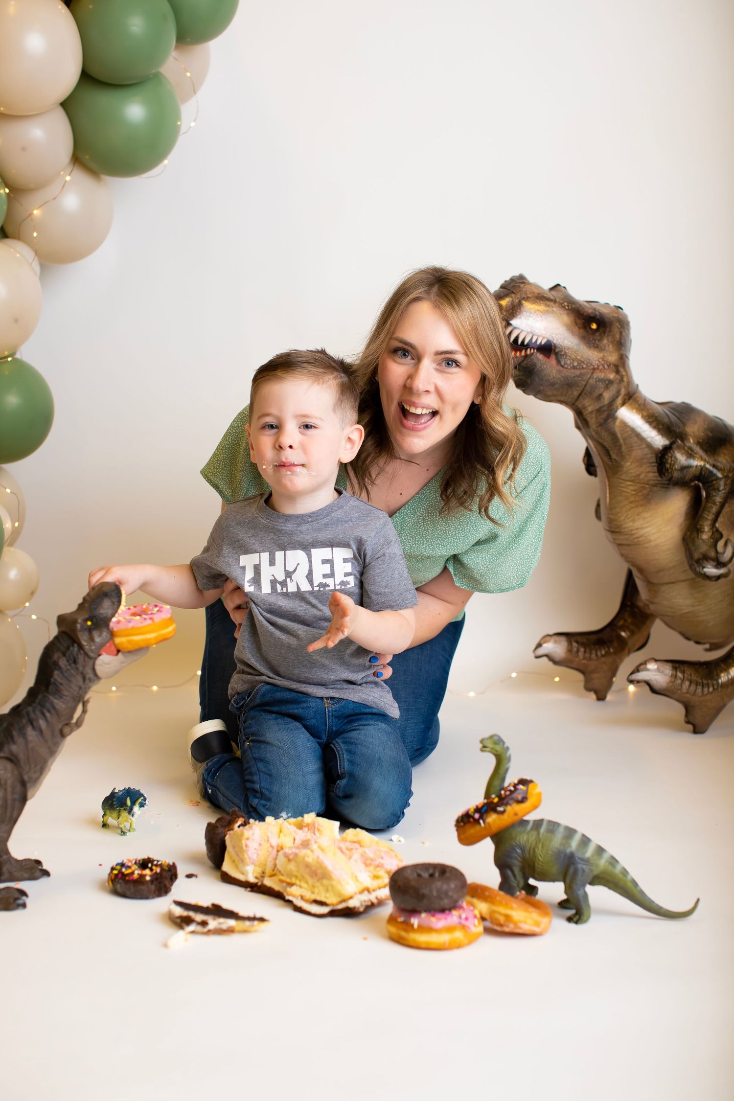 Dinosaur themed celebration with balloon arch, toy dinosaurs, and treats on white background.