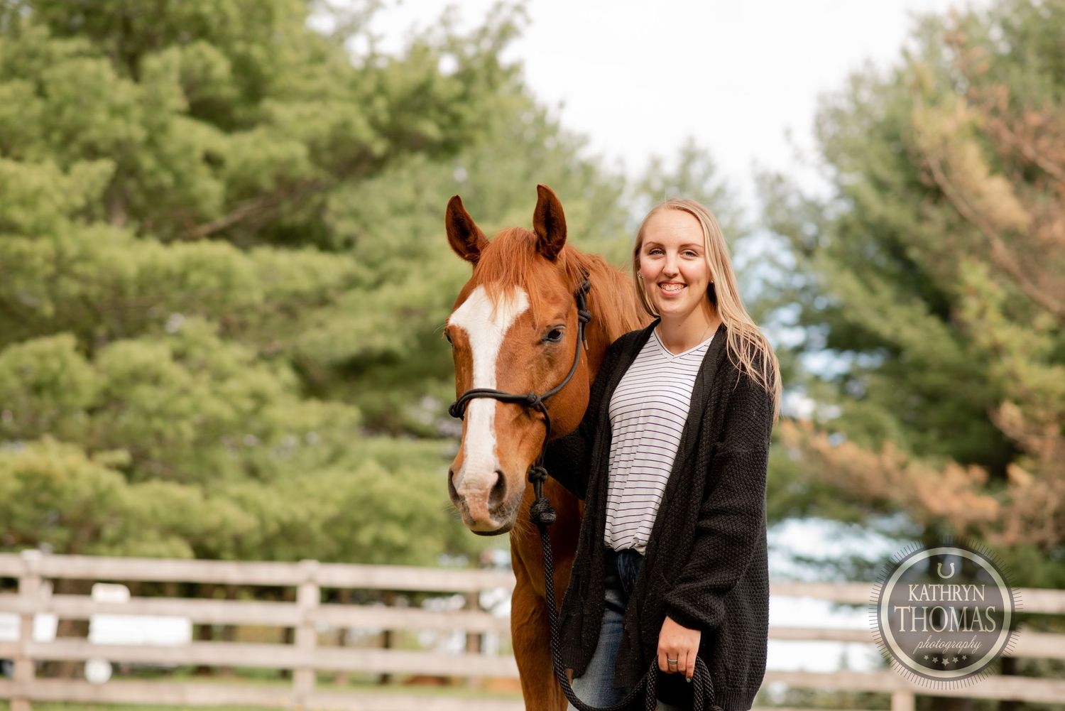 A chestnut horse and companion stand together in a fenced pasture with evergreen trees in the background.