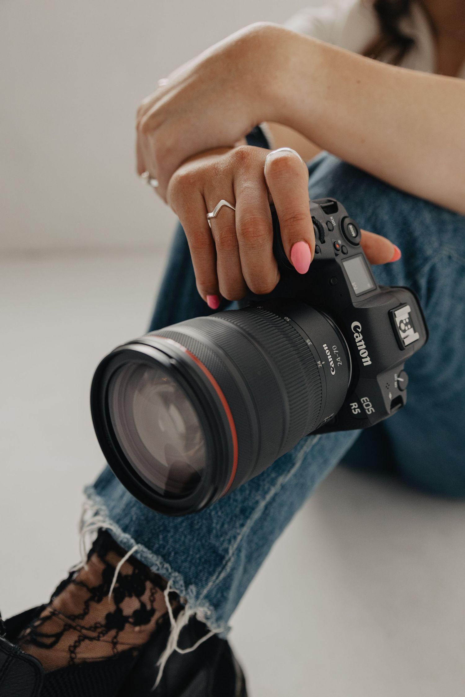 Close-up of hands holding a professional Canon camera with pink nails and ripped jeans.