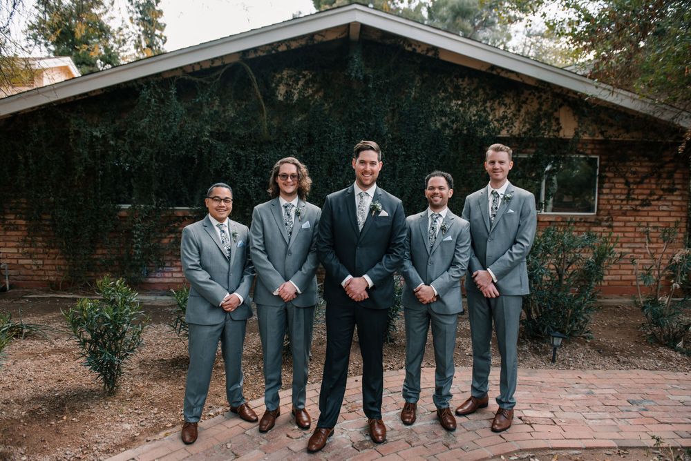 Groomsmen in matching gray suits stand together in front of rustic brick building.