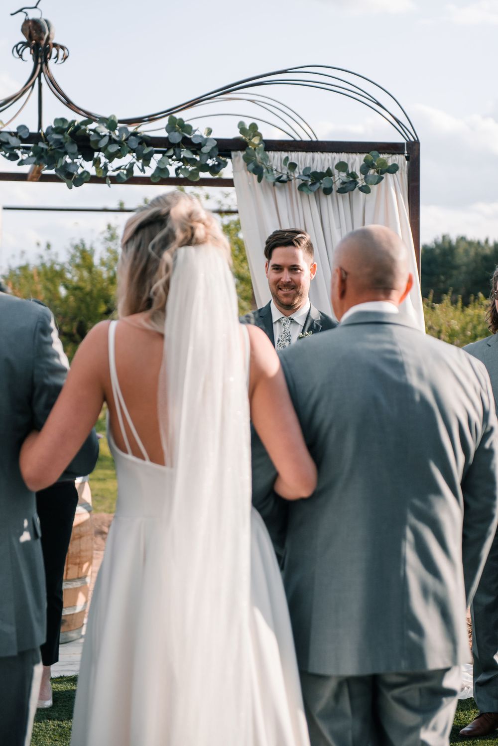 Wedding ceremony moment captured under metal arch decorated with greenery and white fabric.
