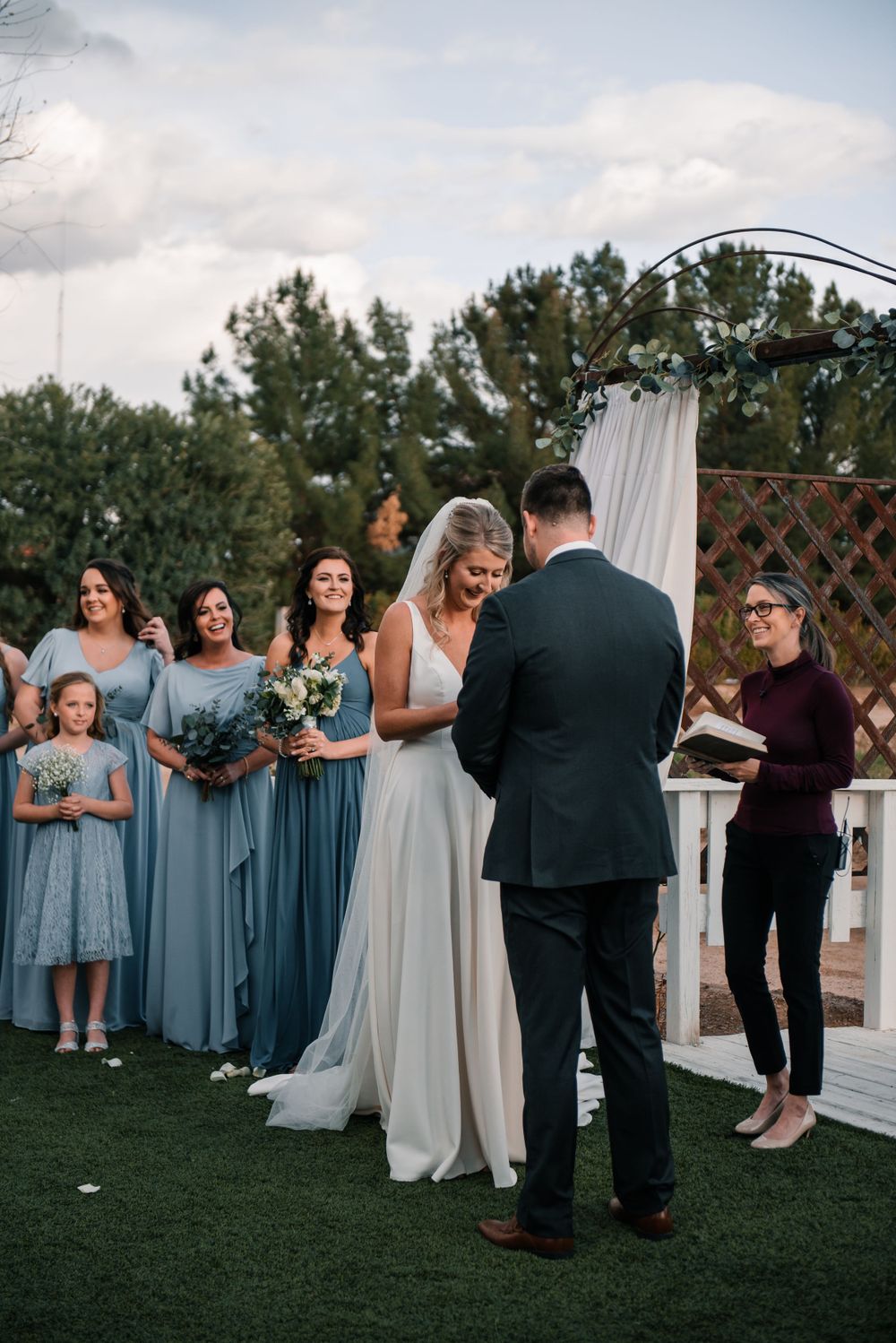 Wedding ceremony with bridesmaids in blue dresses standing beside decorated white arch.