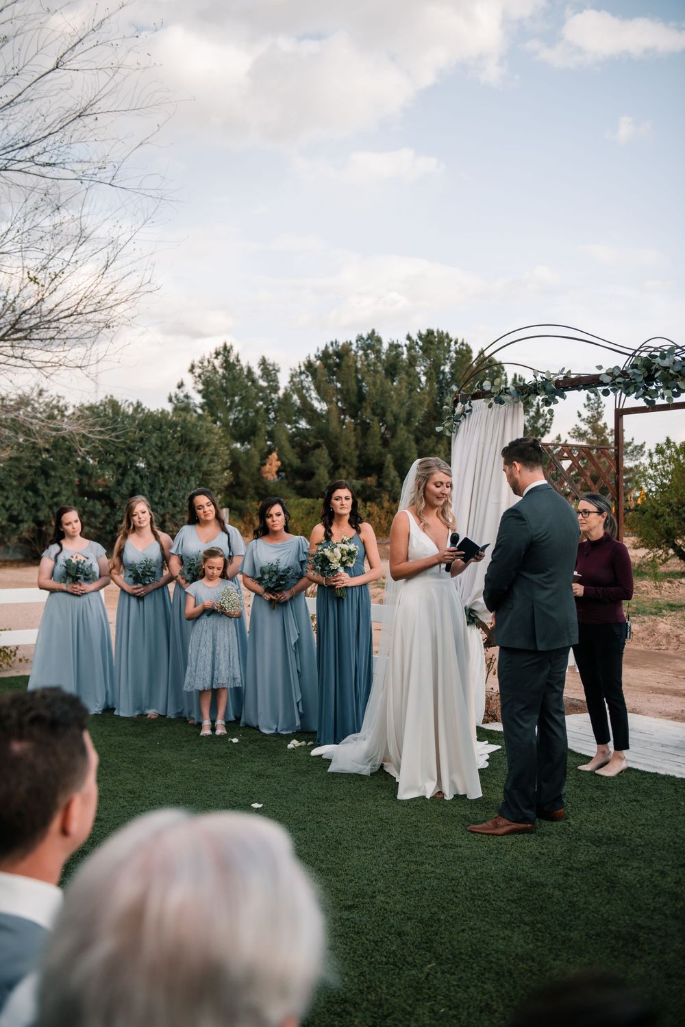 Wedding party in dusty blue bridesmaid dresses stands in a line during outdoor ceremony at sunset.
