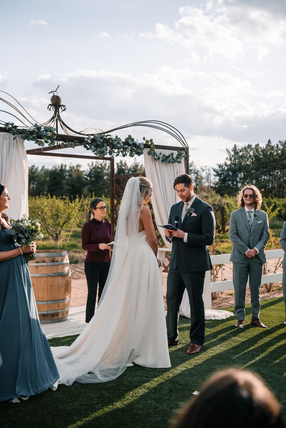 Outdoor wedding ceremony under white draped arch with wedding party in blue and black attire.