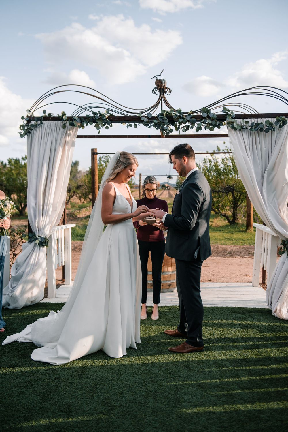 Intimate wedding ceremony moment under white draped arch with guests watching.