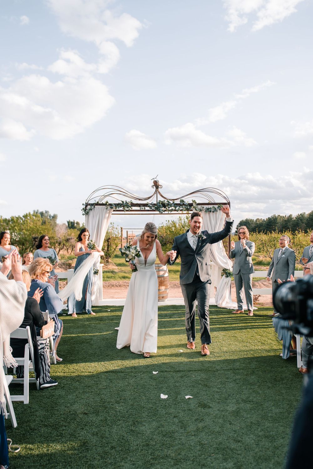 Wedding celebration on green lawn with white chairs and decorated arch in background.