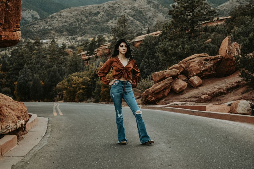 Young woman in rust blouse and flare jeans posing on a mountain road.