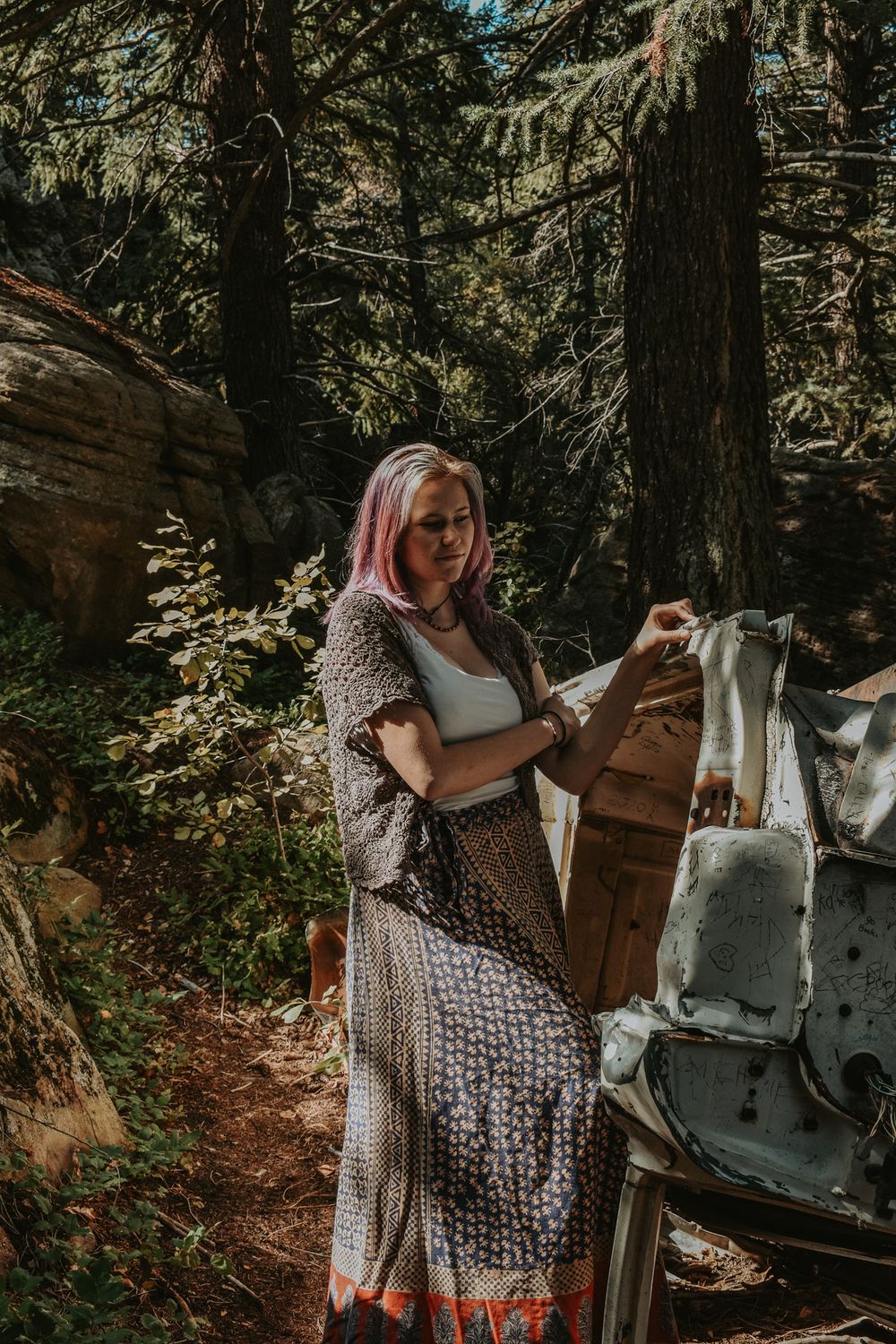 Woman with pink hair in bohemian dress exploring abandoned equipment in a sunlit forest setting.