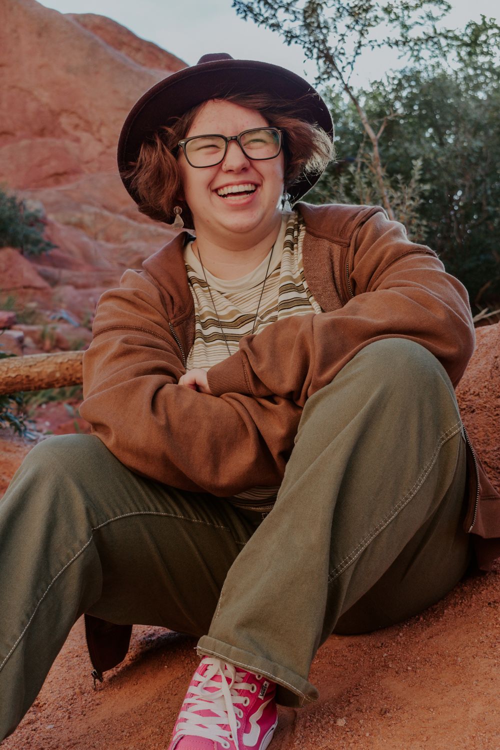Laughing young person in brown jacket and green pants sitting on red rocks outdoors.