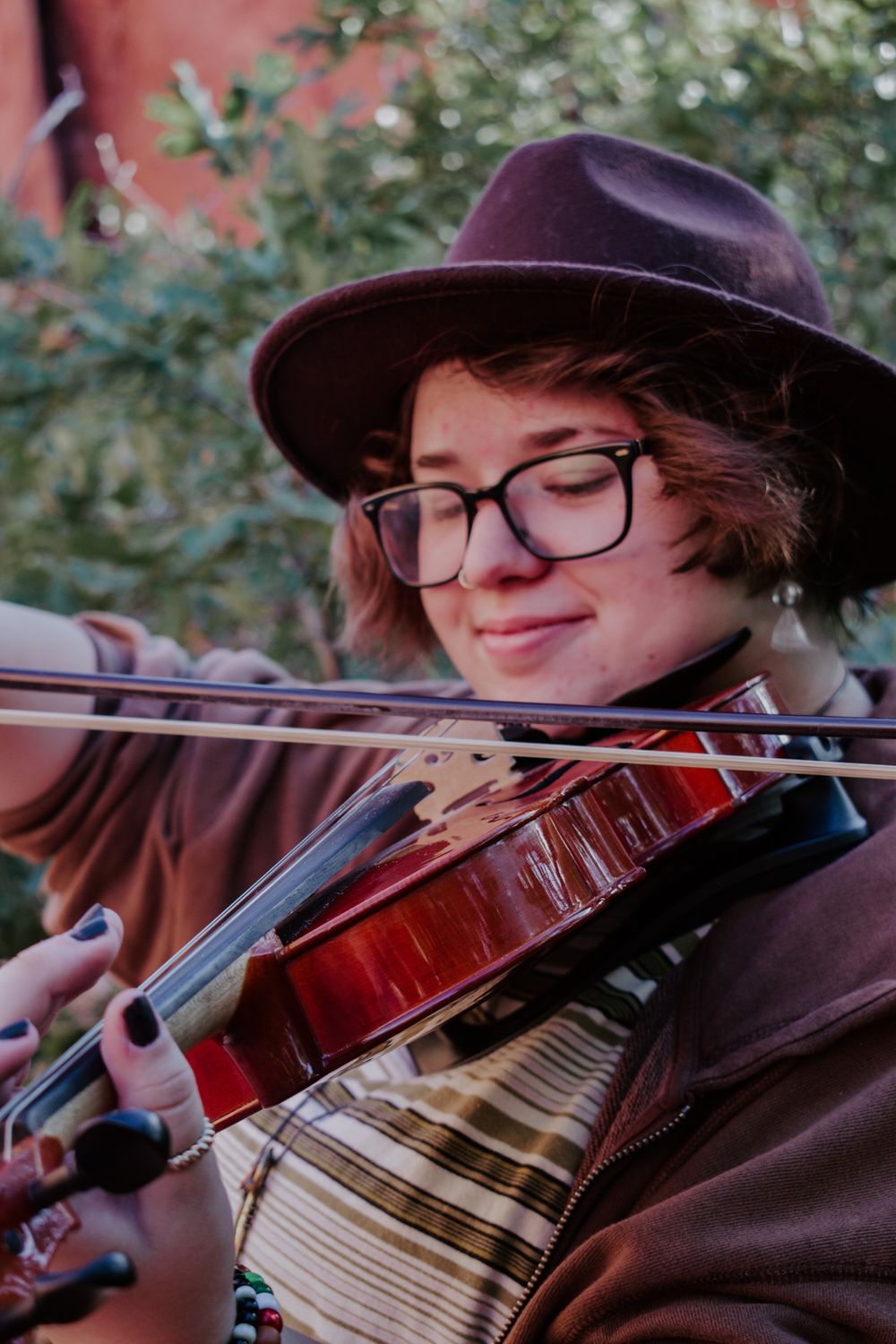 Young musician playing violin outdoors, wearing glasses and a brown wide-brim hat.